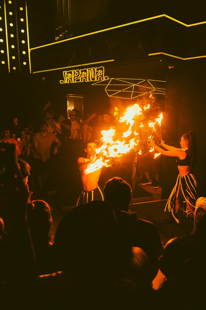 Dos artistas lanzan grandes llamas bajo el neón de JARANA, con el público alrededor, en Bribón del Puerto.